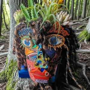 May include: A decorative planter shaped like a human face, adorned with dark brown dreadlocks, beaded blue eyes, and red lips. The head is filled with various green succulents and yellow flowers, featuring a small bee figure on the nose.