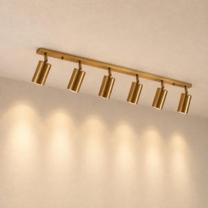 May include: A ceiling-mounted track lighting fixture featuring a long, rectangular brass-toned bar with six adjustable cylindrical spotlights. Each light head is oriented downward, emitting a warm, focused glow onto the surface below.