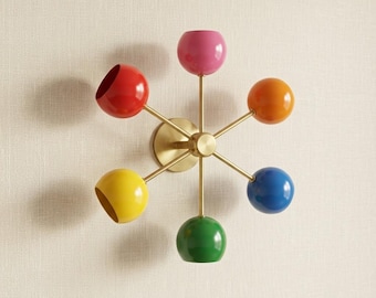 May include: A wall-mounted light fixture featuring a central brass-toned circular base with six radiating arms. Each arm terminates in a spherical shade in a distinct color: pink, orange, blue, green, yellow, and red, arranged in a starburst pattern.