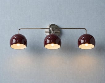 May include: A three-light wall fixture featuring a polished nickel horizontal bar and a circular wall mount. Three glossy, deep burgundy dome-shaped shades are attached to the bar, each housing a single light bulb with a white interior finish.