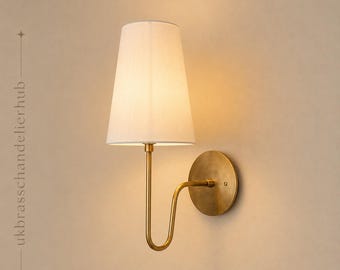 May include: A wall sconce featuring a conical white fabric lampshade attached to a curved brass arm and a circular brass wall plate. The text ukbrasschandelierhub is printed vertically on the left side of the image.