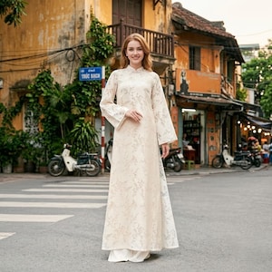 May include: A floor-length, cream-colored dress featuring a high collar, long flared sleeves, and a subtle, repeating floral pattern. The garment is worn over matching wide-leg trousers, creating a layered silhouette with a soft, textured finish.