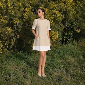 Pode incluir: Um vestido bege de manga curta com decote redondo, detalhes de costura vertical, dois bolsos frontais e uma larga faixa branca na bainha. O vestido tem um corte estruturado e termina acima do joelho.