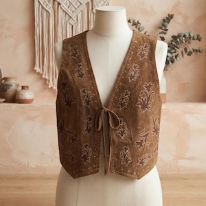 Peut inclure: Un gilet en daim marron orné d'un motif floral brodé répétitif en fil marron foncé. Ce vêtement sans manches présente une encolure en V profonde, une fermeture à nouer sur le devant avec de fines lanières en daim et des surpiqûres décoratives.