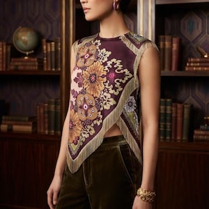 Puede incluir: Una blusa sin mangas con un estampado floral en tonos morado oscuro y dorado, con un dobladillo asimétrico tipo pañuelo adornado con flecos. La prenda se combina con pantalones de terciopelo verde oliva oscuro y un brazalete dorado ancho.