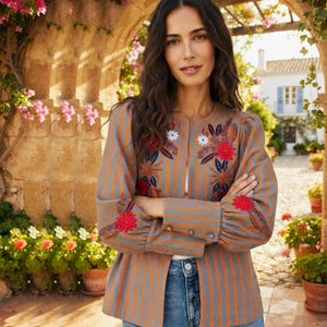 May include: A long-sleeved jacket featuring vertical gray and orange stripes, adorned with embroidered red and white floral patterns on the chest and sleeves. The jacket has a round neckline, buttoned cuffs, and is paired with blue denim jeans.