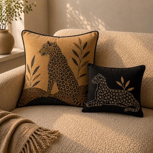 May include: Two square throw pillows featuring leopard illustrations. The larger pillow is tan with a seated leopard and dark leaf motifs. The smaller pillow is black with a reclining leopard and gold-toned leaf accents, both with dark piping.
