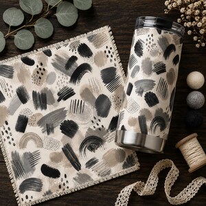 May include: A square fabric napkin with a frayed edge and a cylindrical stainless steel tumbler, both featuring a matching pattern of black, grey, and beige brushstrokes, dots, and arched lines on a cream-coloured background.