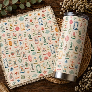 May include: A square fabric napkin with a fringed edge and a cylindrical metal tumbler, both featuring a repeating pattern of colourful mahjong tiles, pink lanterns, and small floral motifs on a cream-coloured background.