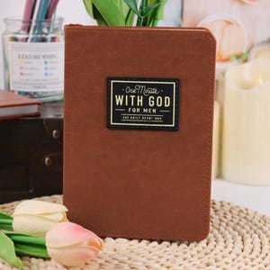Puede incluir: Un diario rectangular de aspecto de cuero marr&oacute;n con una etiqueta rectangular negra en la portada. La etiqueta presenta el texto "One Minute WITH GOD FOR MEN 365 DAILY DEVOTIONS" en letras blancas y doradas con un borde decorativo.