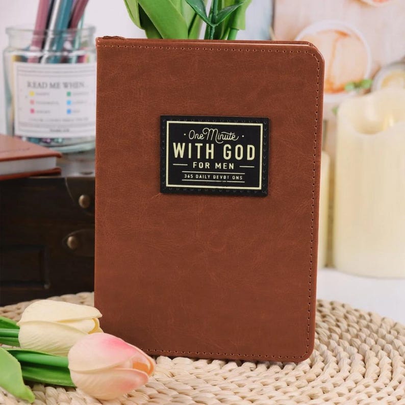 Puede incluir: Un diario rectangular de aspecto de cuero marr&oacute;n con una etiqueta rectangular negra en la portada. La etiqueta presenta el texto "One Minute WITH GOD FOR MEN 365 DAILY DEVOTIONS" en letras blancas y doradas con un borde decorativo.