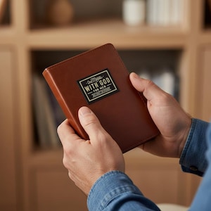 Puede incluir: Un libro encuadernado en cuero marr&oacute;n con una etiqueta rectangular negra en la portada que contiene el texto One Minute WITH GOD FOR MEN. El libro tiene una cubierta lisa de color s&oacute;lido y se sostiene en posici&oacute;n vertical con un dise&ntilde;o sencillo.