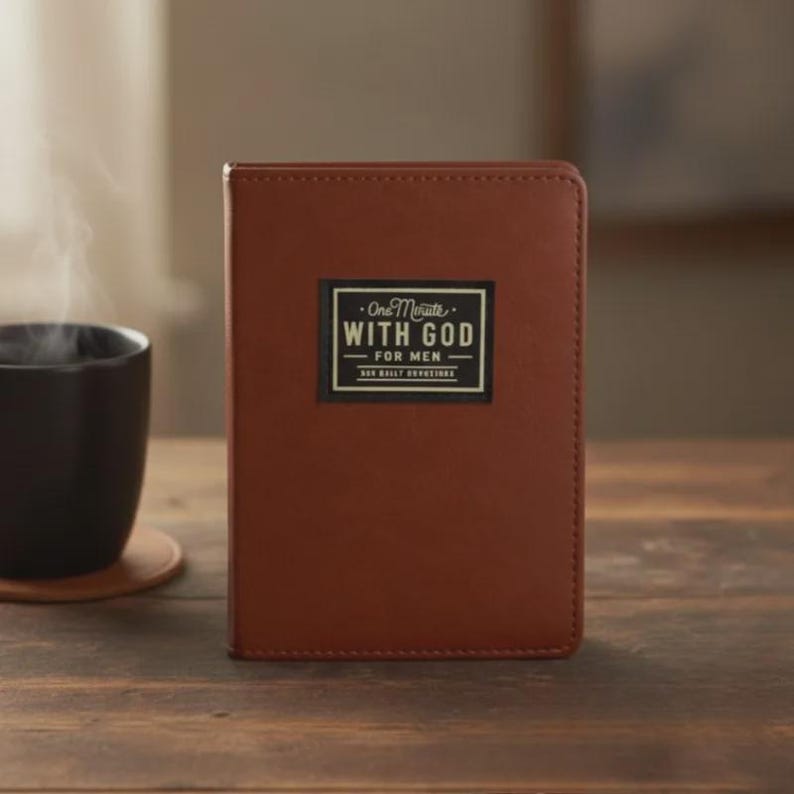 Puede incluir: Un libro encuadernado en cuero marr&oacute;n con una etiqueta negra rectangular en la portada. La etiqueta presenta el texto blanco One Minute WITH GOD FOR MEN. El libro tiene un acabado liso y mate y est&aacute; colocado en posici&oacute;n vertical sobre madera.