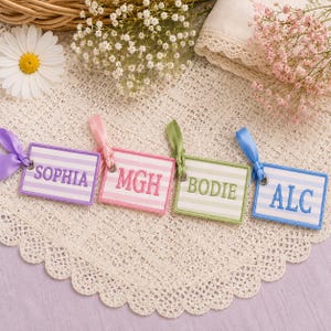 May include: Four rectangular embroidered tags with striped backgrounds and satin ribbons. The tags feature the names or initials SOPHIA, MGH, BODIE, and ALC in matching thread colors. Each tag is finished with a metal eyelet and a colored ribbon.