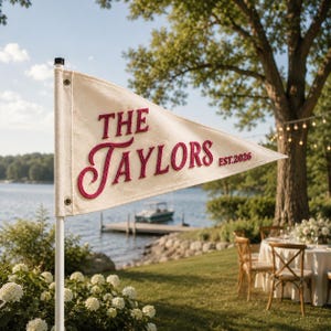 Könnte beinhalten: Ein dreieckiger, cremefarbener Filzwimpel an einem weißen Stab. Die Flagge zeigt den gestickten roten Schriftzug "THE TAYLORS" in einer Mischung aus Block- und Schreibschrift, mit dem kleineren roten Text "EST. 2026" auf der rechten Seite.