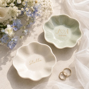 May include: Two flower-shaped ceramic trinket dishes. One is sage green with gold text reading J heart A and 25.08.2016. The other is cream with the gold name Stella. Two gold rings rest nearby. The dishes have a smooth, glossy finish and scalloped edges.
