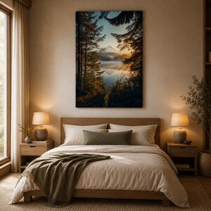 May include: A rectangular wall art print featuring a landscape of a calm lake surrounded by dark pine trees and a snow-capped mountain peak in the distance during sunset, displayed above a bed with beige bedding and a green throw blanket.