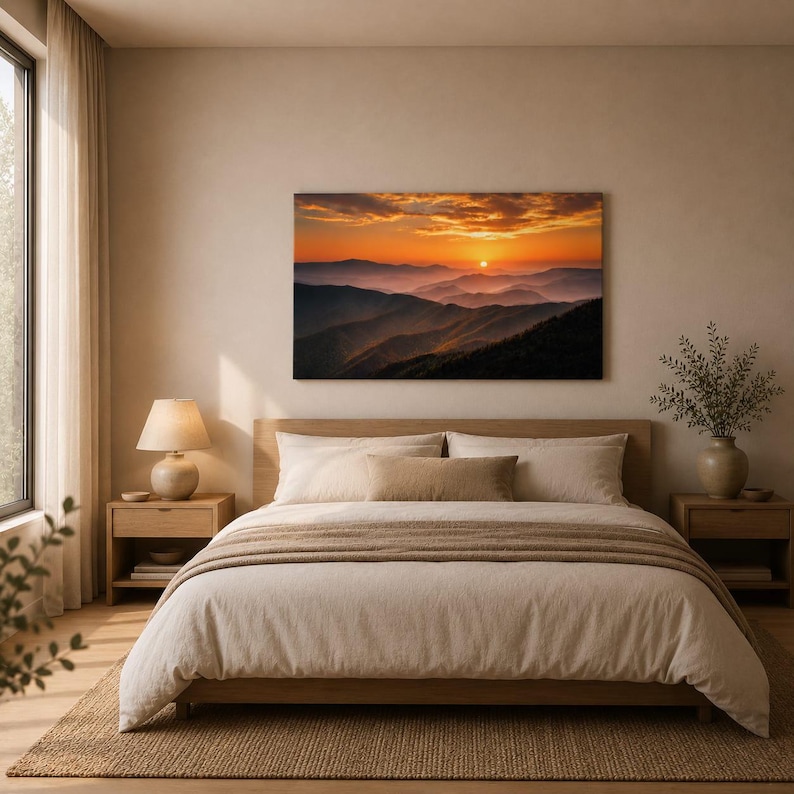 K&ouml;nnte beinhalten: Ein rechteckiger Wandkunstdruck, der ein Gebirge bei Sonnenuntergang mit orangefarbenen und gelben Wolken &uuml;ber geschichteten violetten Bergr&uuml;cken zeigt, platziert &uuml;ber einem Bett mit beiger Bettw&auml;sche und zwei Nachttischen.