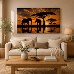 Può includere: Una stampa artistica rettangolare da parete che mostra la silhouette di due elefanti e alberi di acacia contro un vibrante cielo arancione al tramonto, con i loro riflessi visibili nell'acqua. L'opera è esposta sopra un divano beige in salotto.