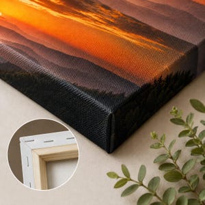 K&ouml;nnte beinhalten: Ein Leinwanddruck mit einem Sonnenuntergang &uuml;ber geschichteten Bergk&auml;mmen mit strukturierter Oberfl&auml;che. Der Einschub zeigt die R&uuml;ckseite der Leinwand und offenbart einen hellen Holzrahmen mit Metallklammern, die den Stoff am Holz befestigen.