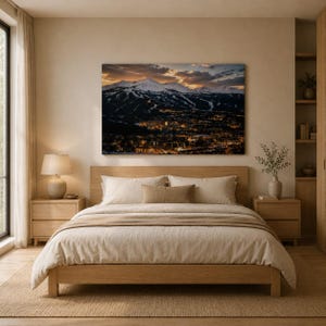 May include: A bedroom featuring a wooden bed frame with beige bedding, two wooden nightstands, and a table lamp. Above the bed hangs a large rectangular wall art print depicting a snow-capped mountain range at dusk overlooking a glowing town.