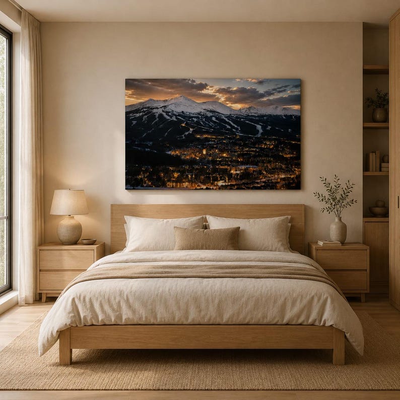 May include: A bedroom featuring a wooden bed frame with beige bedding, two wooden nightstands, and a table lamp. Above the bed hangs a large rectangular wall art print depicting a snow-capped mountain range at dusk overlooking a glowing town.
