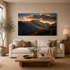 Könnte beinhalten: Ein großes rechteckiges Wandbild mit einer schneebedeckten Bergkette bei Sonnenuntergang, goldenem Licht auf den Gipfeln und dunklen Wolken, das in einem Wohnzimmer über einem beigen Sofa mit Kissen und einem hölzernen Couchtisch hängt.