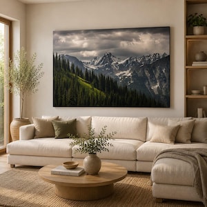 Könnte beinhalten: Ein großes rechteckiges Wandbild, das eine dramatische Bergkette mit schneebedeckten Gipfeln, dunklen Nadelwäldern und einem bewölkten Himmel zeigt, angebracht über einem beigen Ecksofa mit einem runden Couchtisch aus Holz und einer Pflanze.
