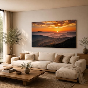K&ouml;nnte beinhalten: Ein gro&szlig;es rechteckiges Wandbild, das einen Sonnenuntergang &uuml;ber geschichteten Gebirgsz&uuml;gen in Orange-, Gold- und tiefen Violettt&ouml;nen zeigt. Das Kunstwerk ist &uuml;ber einem beigen Ecksofa mit mehreren Zierkissen in einem Wohnzimmer angebracht.