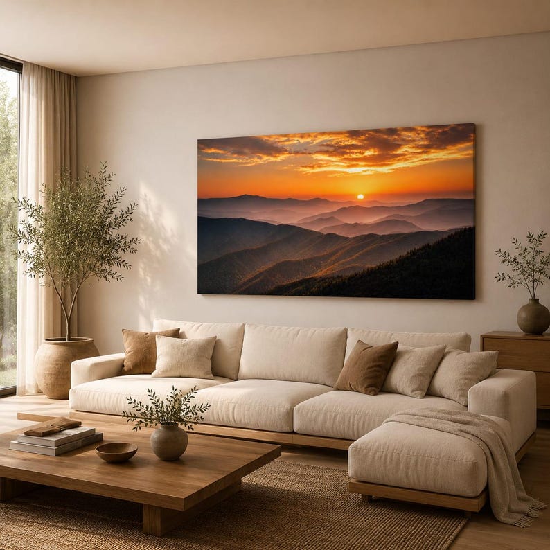 K&ouml;nnte beinhalten: Ein gro&szlig;es rechteckiges Wandbild, das einen Sonnenuntergang &uuml;ber geschichteten Gebirgsz&uuml;gen in Orange-, Gold- und tiefen Violettt&ouml;nen zeigt. Das Kunstwerk ist &uuml;ber einem beigen Ecksofa mit mehreren Zierkissen in einem Wohnzimmer angebracht.