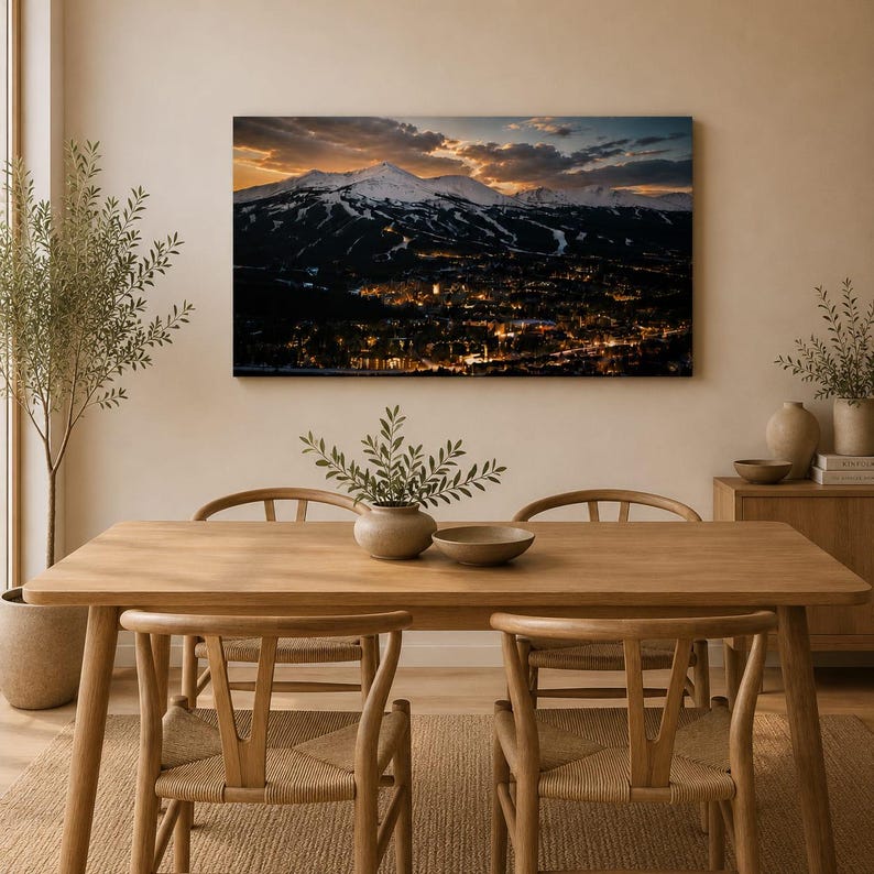 May include: A rectangular wall art print featuring a panoramic landscape of snow-capped mountains at sunset overlooking a town with glowing lights. The print is displayed in a dining room above a wooden table with chairs and a small potted plant.