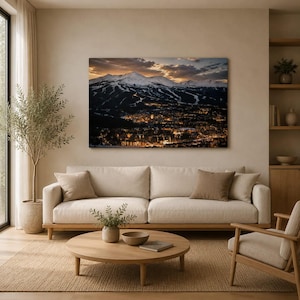 Könnte beinhalten: Ein rechteckiger Wandkunstdruck, der ein schneebedecktes Gebirge bei Sonnenuntergang über einer Stadt zeigt, die nachts von warmem goldenem Licht beleuchtet wird. Das Landschaftsfoto hängt in einem Wohnzimmer über einem beigen Sofa.
