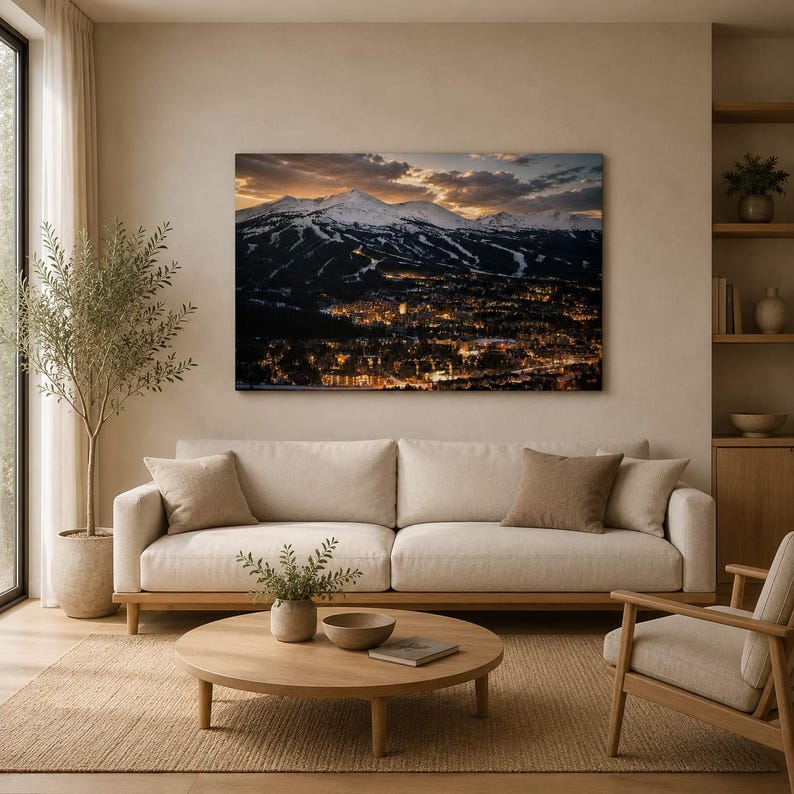 May include: A rectangular wall art print depicting a snow-capped mountain range at sunset overlooking a town illuminated by warm golden lights at night. The landscape photograph is displayed in a living room above a beige sofa and wooden coffee table.