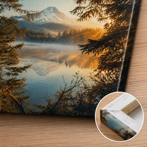 May include: A rectangular canvas print featuring a landscape of a snow-capped mountain reflected in a calm lake at sunrise, framed by dark pine tree silhouettes. The inset shows the back corner of the canvas with staples and a wooden frame.