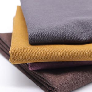 May include: A stack of four folded fabric swatches featuring a distinct diagonal twill weave texture. The colors from top to bottom are charcoal gray, mustard yellow, deep plum purple, and a dark chocolate brown, all neatly layered on top of each other.