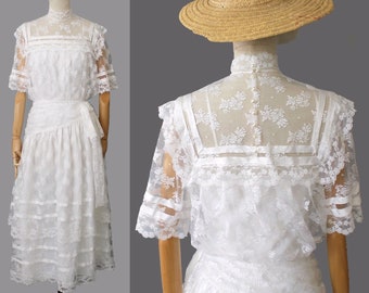 Op de afbeelding: Witte kantenjurk met een hoge hals, korte mouwen en een gelaagde rok. De jurk heeft een ceintuur die in de taille wordt geknoopt. De jurk is op een mannequin.