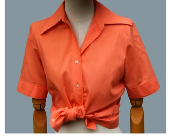 Puede incluir: Una camisa de manga corta de color coral con botones y cuello. La camisa está anudada a la cintura y está en un maniquí.