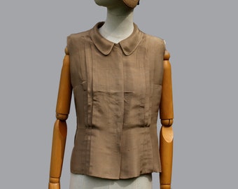 May include: A brown sleeveless blouse with a white collar and a pleated front. The blouse is on a wooden mannequin wearing a straw hat with a black ribbon.