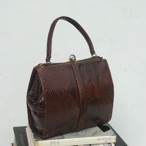 May include: A brown snakeskin handbag with a top clasp closure and a single handle. The bag is sitting on a stack of books.