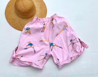 Puede incluir: Shorts de algodón rosa con un detalle de lazo en los lados. Los shorts presentan un patrón repetido de figuras en varias poses que llevan ropa colorida.