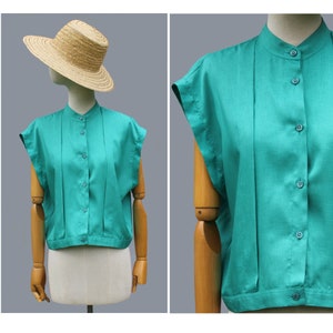 May include: Teal-colored, short-sleeved blouse with a mandarin collar and button-down front. The blouse features pleats down the front and is displayed on a mannequin. A straw hat is on the mannequin's head.
