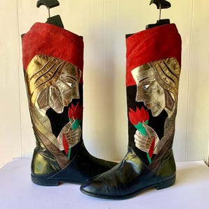 Puede incluir: Un par de botas altas con una sección inferior de cuero negro y un puño superior de ante rojo. La caña muestra una aplicación de metal dorado de un rostro de perfil que sostiene una flor roja con hojas verdes sobre un fondo de ante negro.