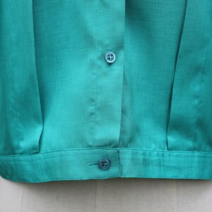 May include: Close-up of a teal-colored blouse. The short-sleeved top features a button-down front with two visible buttons and a textured fabric. The blouse is a solid color and appears to be made of a lightweight material.