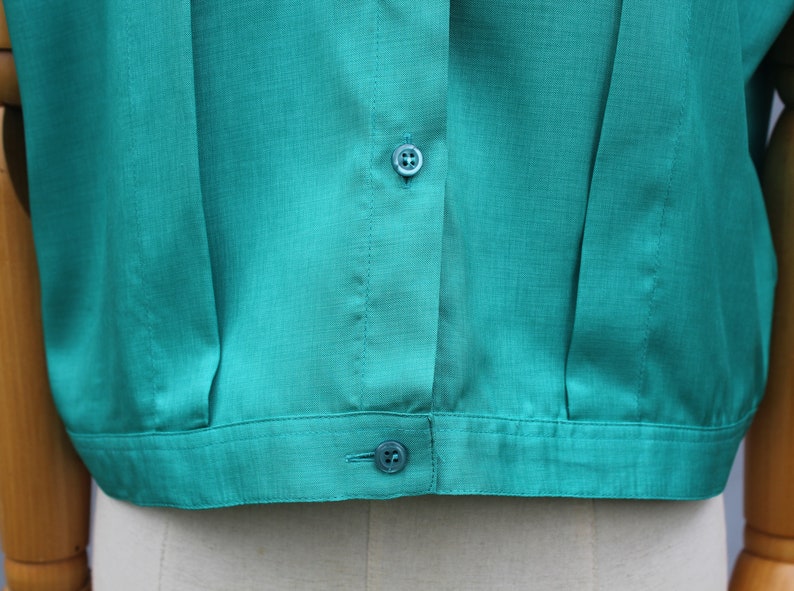 May include: Close-up of a teal-colored blouse. The short-sleeved top features a button-down front with two visible buttons and a textured fabric. The blouse is a solid color and appears to be made of a lightweight material.