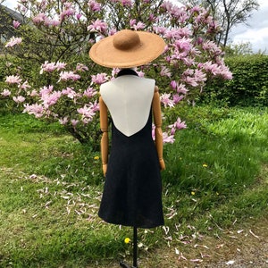 May include: A black sleeveless dress with a white V-shaped panel in the back. The dress is on a mannequin wearing a wide-brimmed straw hat. The mannequin is standing in front of a flowering magnolia tree.