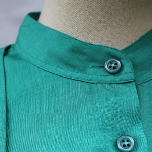 May include: Close-up of a teal-colored shirt with a mandarin collar. The shirt features two matching teal buttons and a visible seam detail. The fabric has a subtle texture, and the collar is neatly stitched.