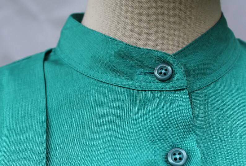 May include: Close-up of a teal-colored shirt with a mandarin collar. The shirt features two matching teal buttons and a visible seam detail. The fabric has a subtle texture, and the collar is neatly stitched.