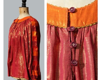 May include: A long-sleeved blouse with a metallic red fabric and gold accents. The blouse features an orange collar and sleeve accents, with three dark purple buttons down the front. The fabric has a vertical striped pattern.