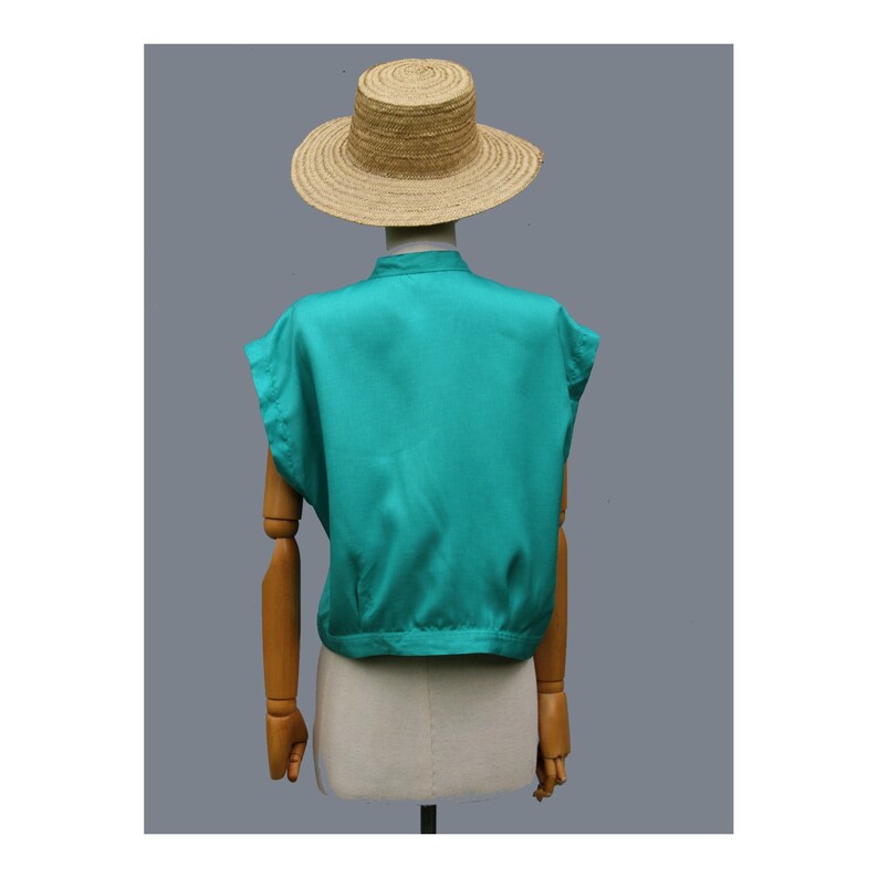 May include: A teal-colored, short-sleeved top with a high collar and a straw hat. The top is made of a shiny fabric and has a gathered hem. The hat is a light brown color and has a wide brim. The clothing is displayed on a mannequin.