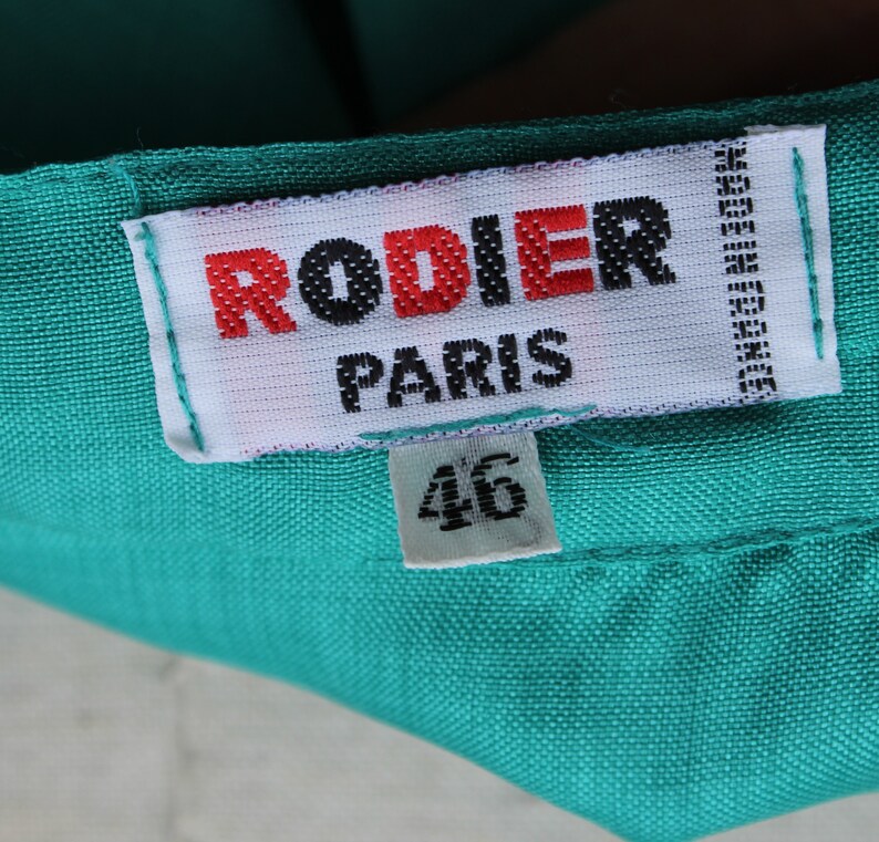 May include: A close-up of a teal fabric with a white label. The label reads "RODIER PARIS" in red and black text, with "46" below. The label also says "MADE IN FRANCE".